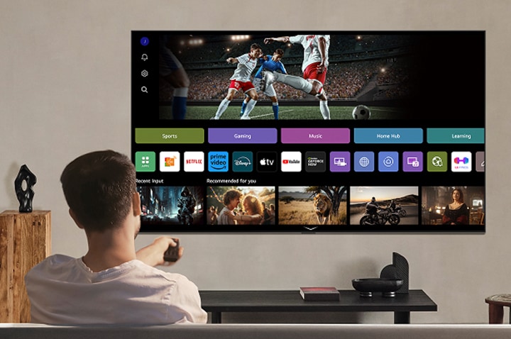 En person på en soffa som håller ut en fjärrkontroll. En väggmonterad LG AI TV med LG webOS på skärmen.