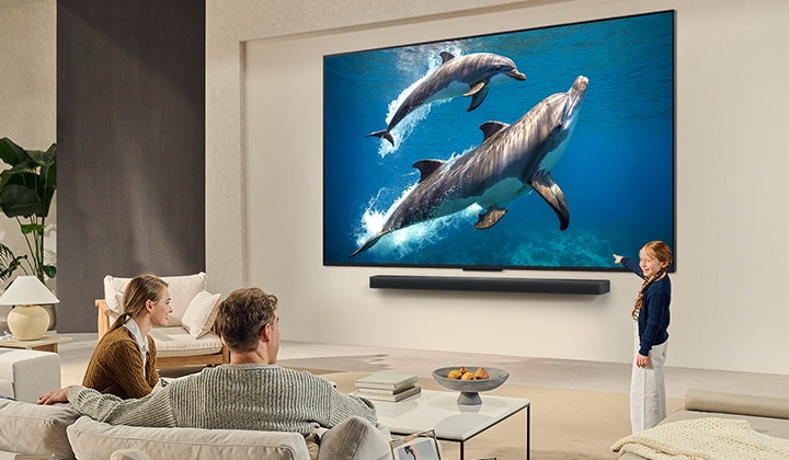 En familj sitter i en soffa vända mot en LG QNED TV monterad på en vägg med en liten flicka som pekar på en skärm som visar två delfiner.