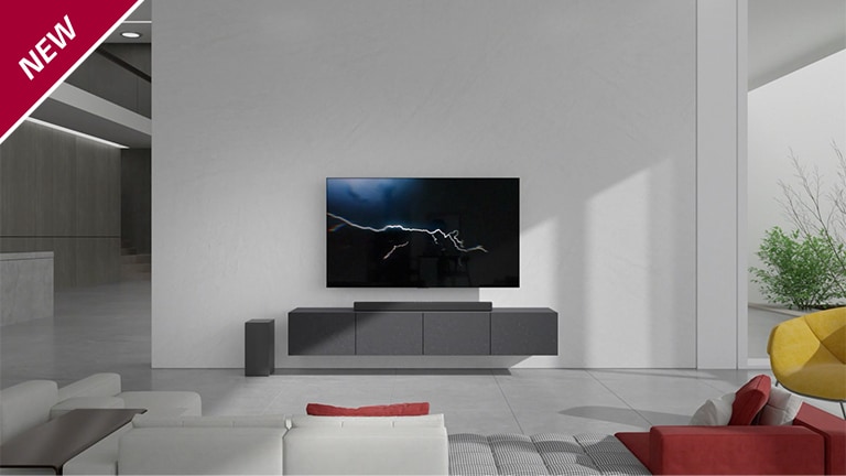 Soundbaren står på ett grått skåp under en TV som hänger på väggen i ett vardagsrum. En trådlös subwoofer står på golvet på vänster sida och solljuset kommer in från bildens högra sida. En vit och röd lång soffa står vänd mot TV-apparaten och soundbaren. NEW-märket visas i övre vänstra hörnet.