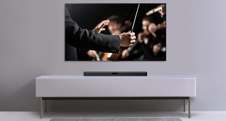 En TV visas på en grå vägg med en LG Soundbar under den på en grå hylla. TV:n visar en dirigent som dirigerar en orkester.