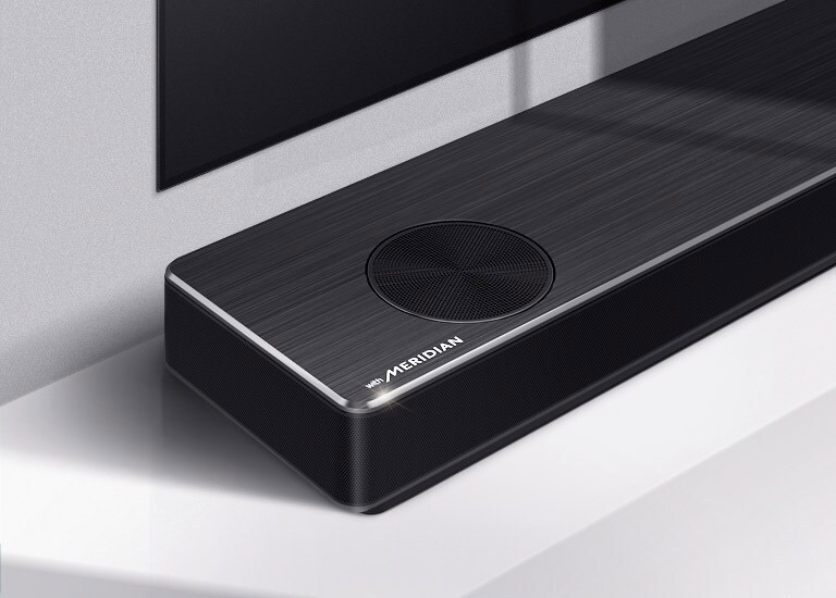 Närbild av LG Soundbars vänstra sida med Meridians logotyp i nedre vänstra hörnet. Nedre vänstersida av TV:n är också synlig.