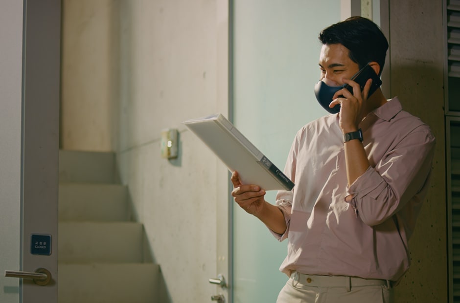 An Asian man is holding a document and talking on the phone, with a black LG PuriCare™ Wearable Air Purifier on.