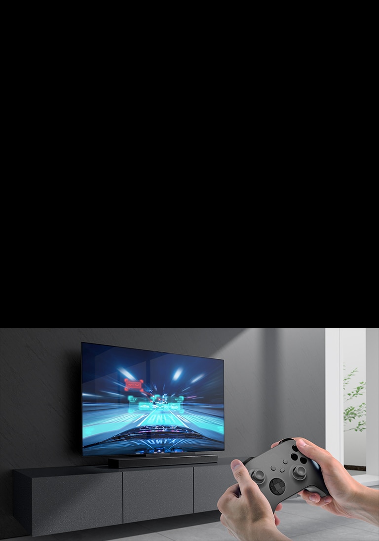 The sound bar is placed on the cabinet and racing game scene is shown on the TV coneected to the sound bar. A game console is on the bottom right side of the picture hold by two hands.