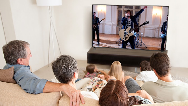 Family of seven gathered in the living room,  watching a movie. TV screen shows a band performing.