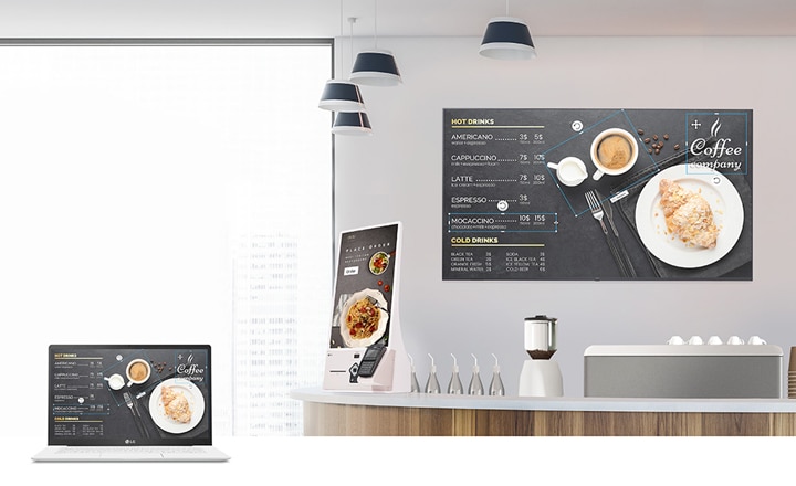 The café's menu board content is being designed using a laptop. Once created, this menu board content is distributed and presented on the signage installed within the cafe.