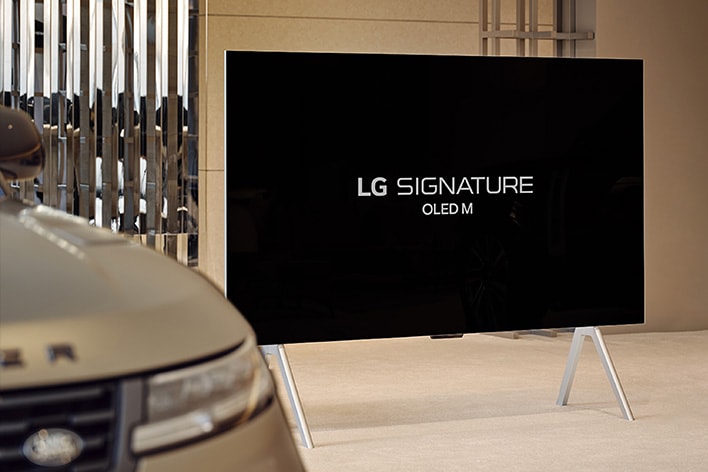 LG SIGNATURE OLED M4 บนขาตั้งที่ Range Rover House ข้าง Range Rover Sport SV และมีปุ่มเล่นที่ด้านล่างของภาพ