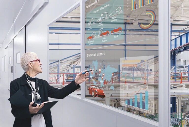 ผู้หญิงคนหนึ่งกำลังดูข้อมูลการทำงานของเธอผ่านจอแสดงผลแบบโปร่งใสที่ติดตั้งอยู่ในหน้าต่างสำนักงาน