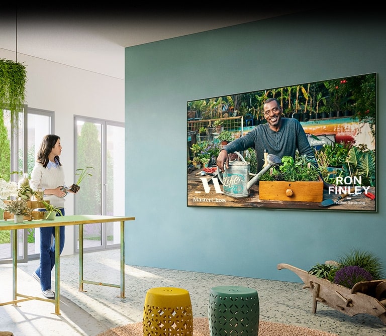 Duvardaki geniş TV'de "Master Class" bahçıvanlık dersi ekranı gösterilir. Televizyonun yanındaki bir masada durarak saksılar ve çiçek makasıyla bahçıvanlık dersi alan kadın.