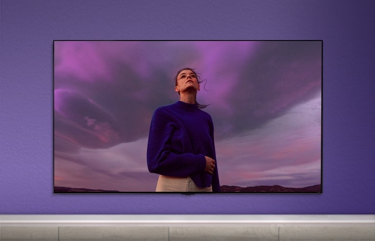 Mor bir duvara yerleştirilmiş QNED TV'nin ekranında mor gömlek giymiş bir kadın.