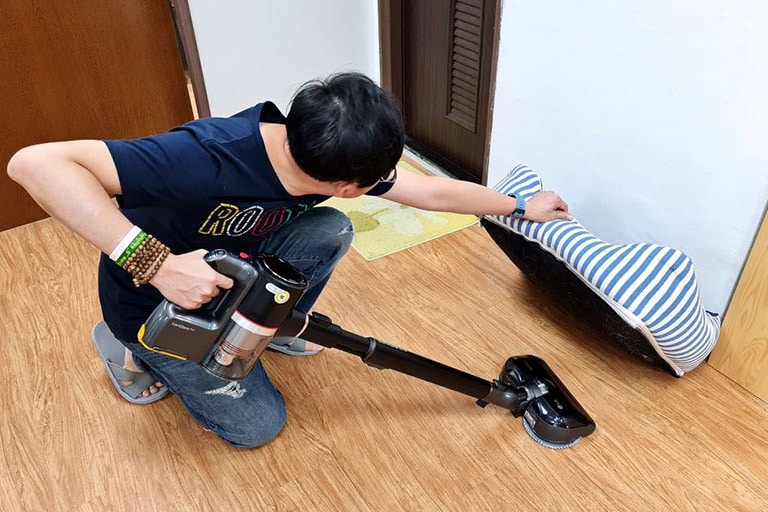 A man crouches down to lift up a dog bed and vacuums beneath it.