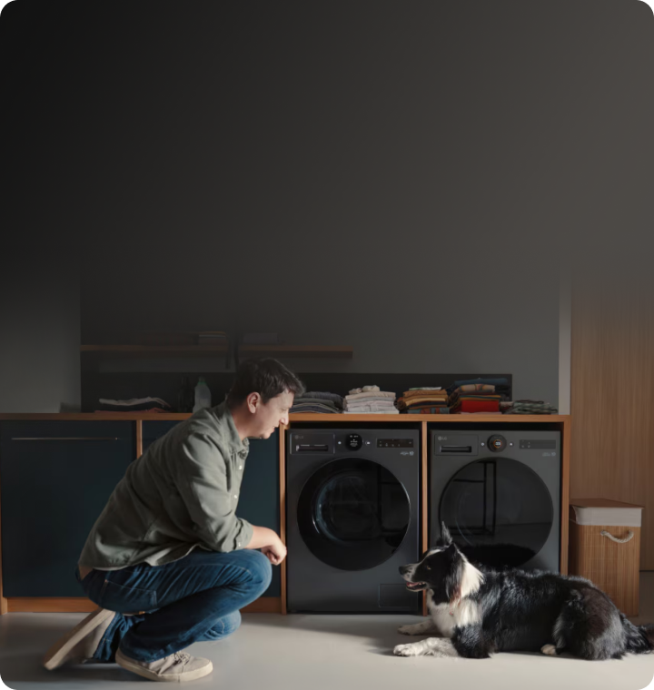 In a home laundry room, a man and a dog are facing each other, with two matte black LG washing machines visible directly behind them.