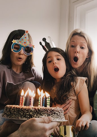 Image of two adult women and a young girl wearing a birthday hat on their head and blowing out the candles on the cake.