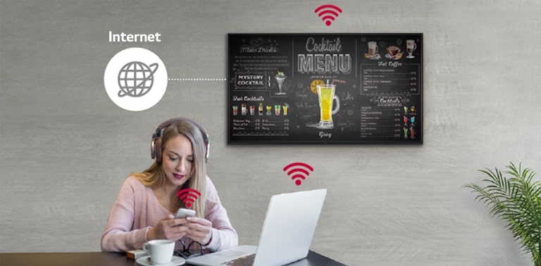 The UR3J series is on the wall and a woman is using a personal PC and mobile phone. This image shows that the signage can be connected wirelessly to the PC and mobile phone which she is holding.