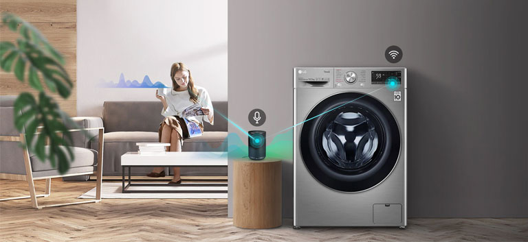 A woman sitting on the couch and drinking tea is giving orders to her washing machine through her AI speaker.