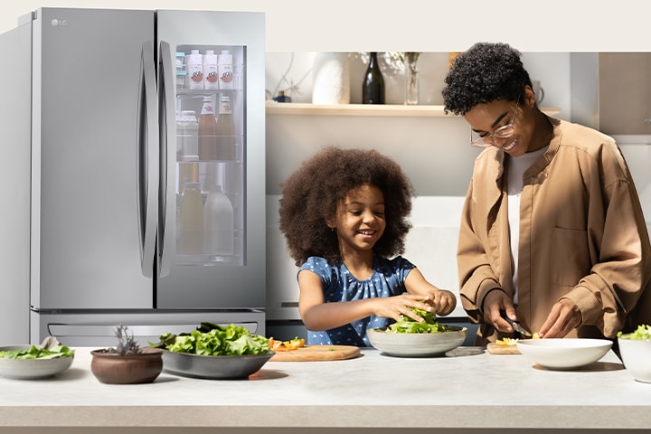 An adult and a child are cooking in a kitchen with a lighted refrigerator