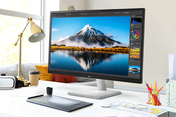 On the desk, there is an ultrafine monitor displaying Photoshop work. Next to the monitor, there is a desk lamp, a mouse, paper, and coloured pencils.