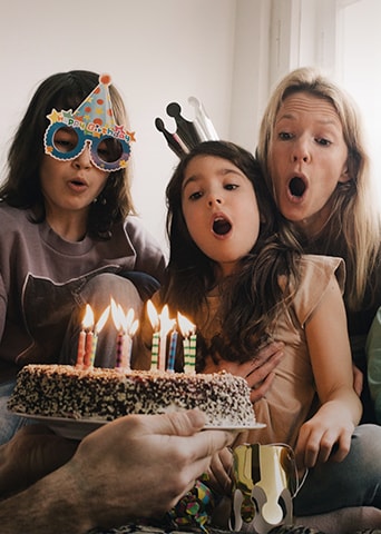 Image of two adult women and a young girl wearing a birthday hat on their head and blowing out the candles on the cake.