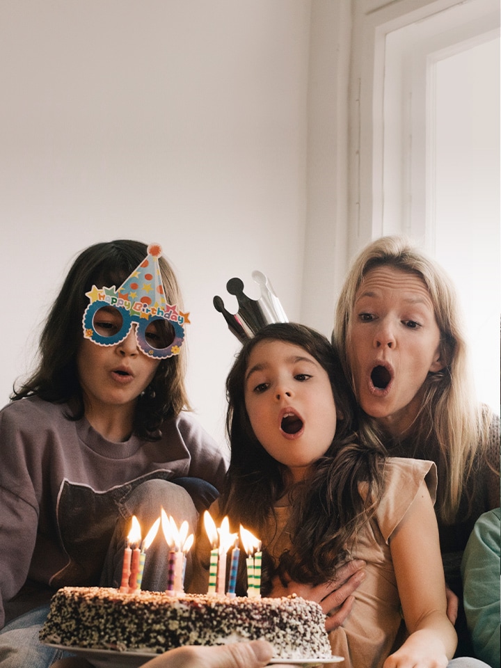 Image of two adult women and a young girl wearing a birthday hat on their head and blowing out the candles on the cake.