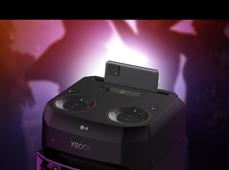 A close-up view of the top of LG XL2S, highlighting on the phone cradle. A smartphone is placed on the phone cradle. Behind, there are silhouettes of people dancing.