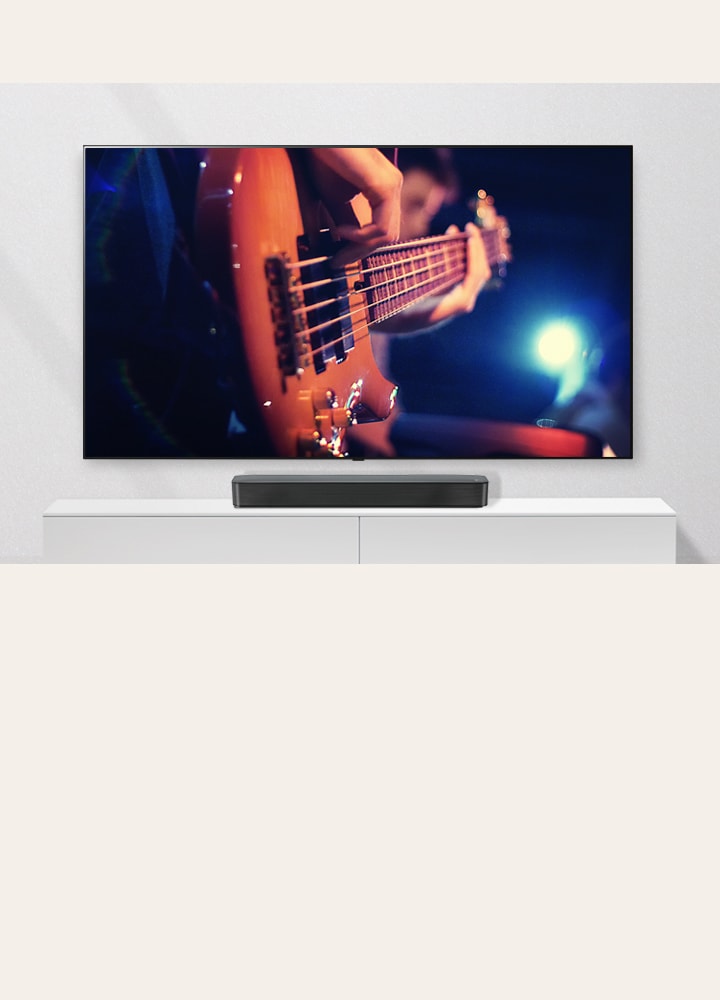 The TV is attached to the wall, and the sound bar is on a white shelf. TV showing a man plays guitar.	