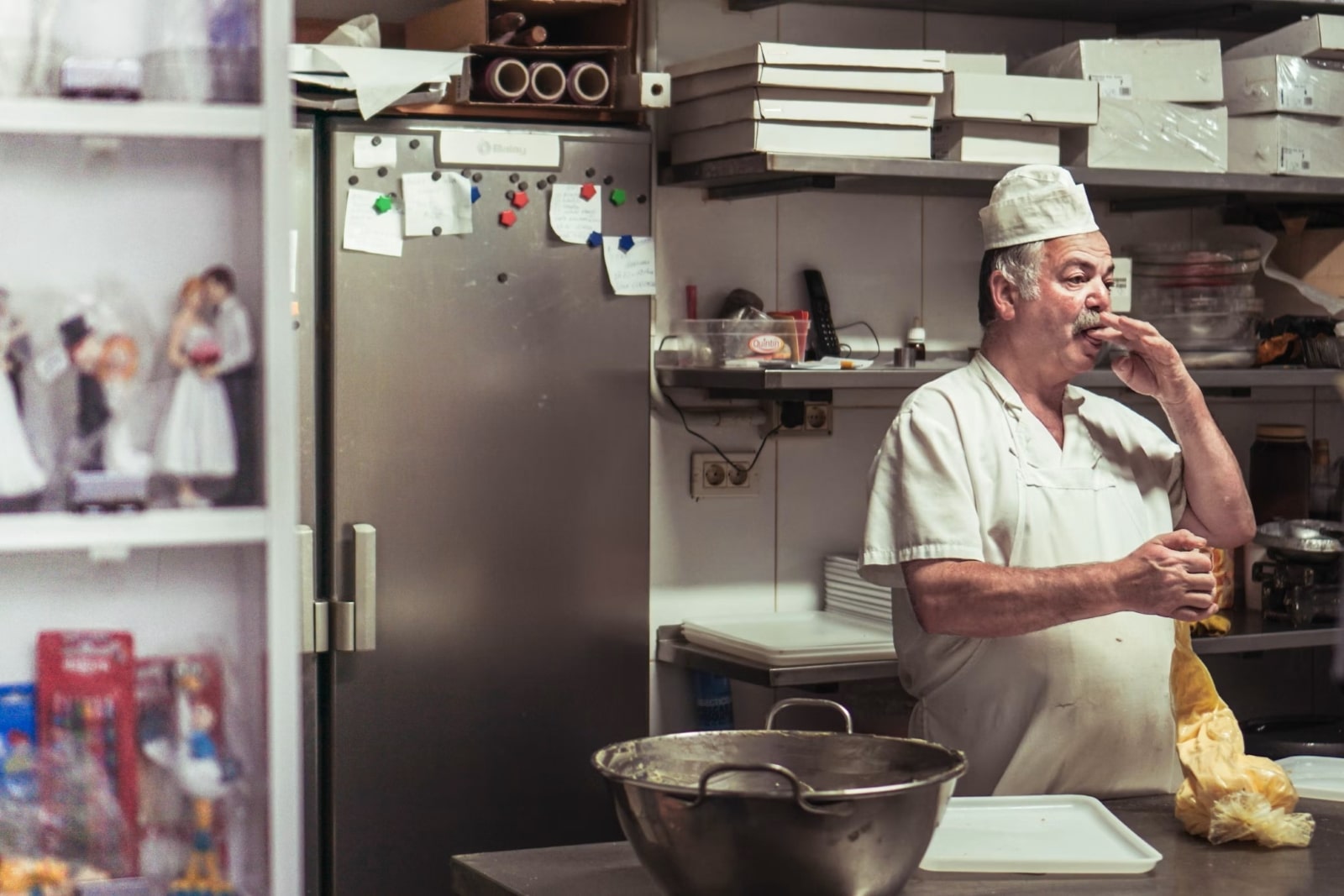 A baker in a white uniform in a kitchen. The background includes a fridge with notes, shelves with boxes, and various baking tools.