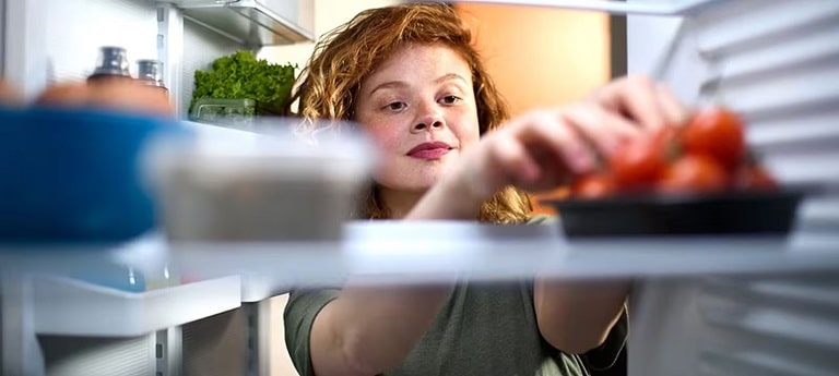 The view is from inside the fridge, looking out. A person is reaching inside the fridge for tomatoes.