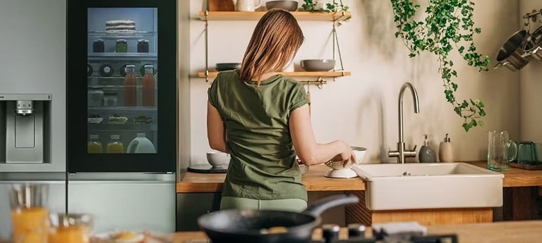 A person is standing in the kitchen and organising items on the kitchen counter. There is a refrigerator next to the person with a transparent panel, with juices and pastries inside. 