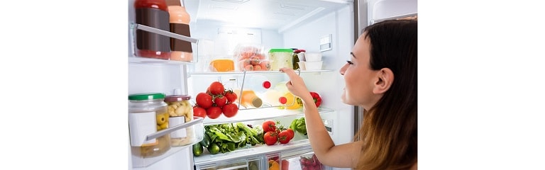  A woman is looking into a fully-stocked fridge filled with tomatoes, vegetables, juices, and more. 