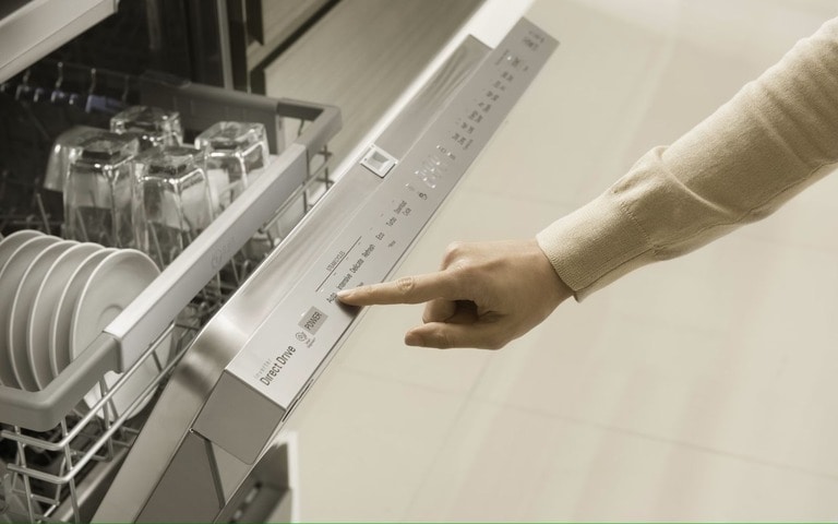 A close-up image of a person's hand pressing a button on the control panel of a stainless steel dishwasher. The dishwasher door is slightly open, showing clean dishes inside.