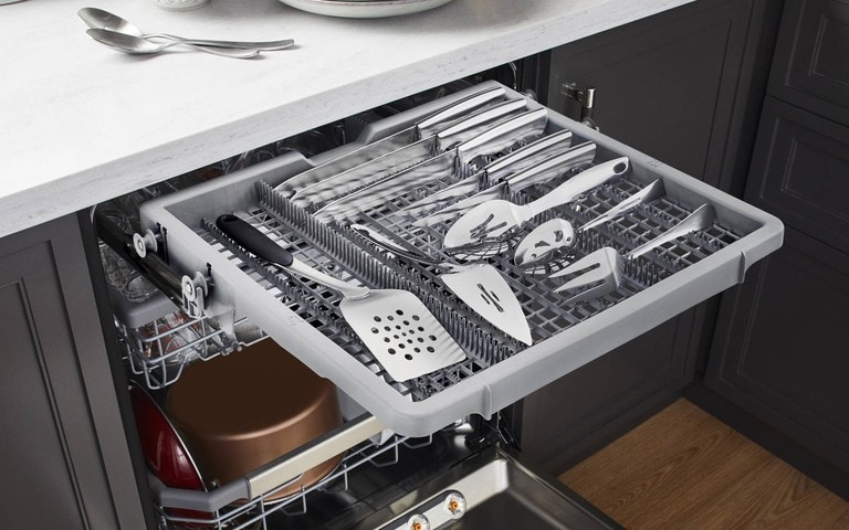 An open LG dishwasher with a dedicated top rack for utensils, neatly holding knives, spatulas, and kitchen tools, set in a modern kitchen with dark cabinetry and a marble countertop