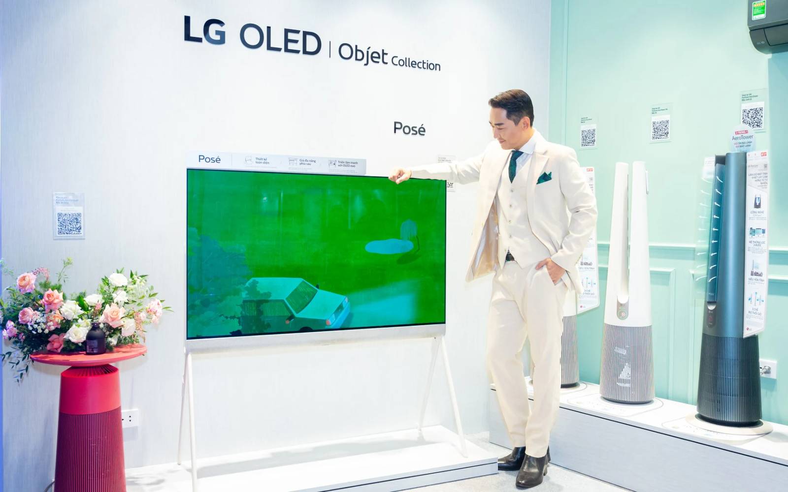 A man in a stylish white suit poses beside an LG OLED Objet Collection Posé TV in a showroom, surrounded by modern appliances and decorative elements