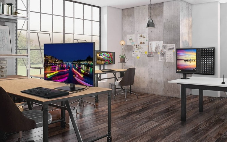 A modern office space featuring multiple workstations with large computer monitors on wooden and white desks, bright natural light streaming through large windows, and a contemporary interior with industrial and minimalist design elements.