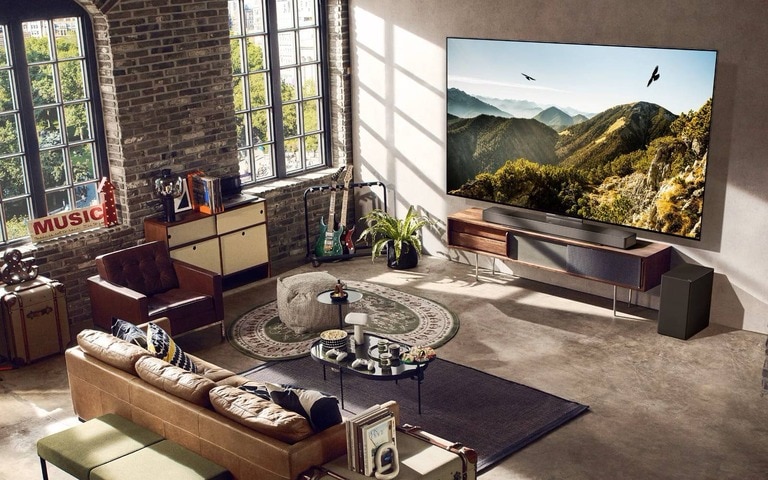 A stylish, spacious loft-style living room featuring a large flat-screen TV mounted on the wall, displaying a scenic mountain view. The room includes exposed brick walls, large industrial-style windows, a leather sofa, musical instruments, and various decor elements that create a warm, artistic atmosphere.