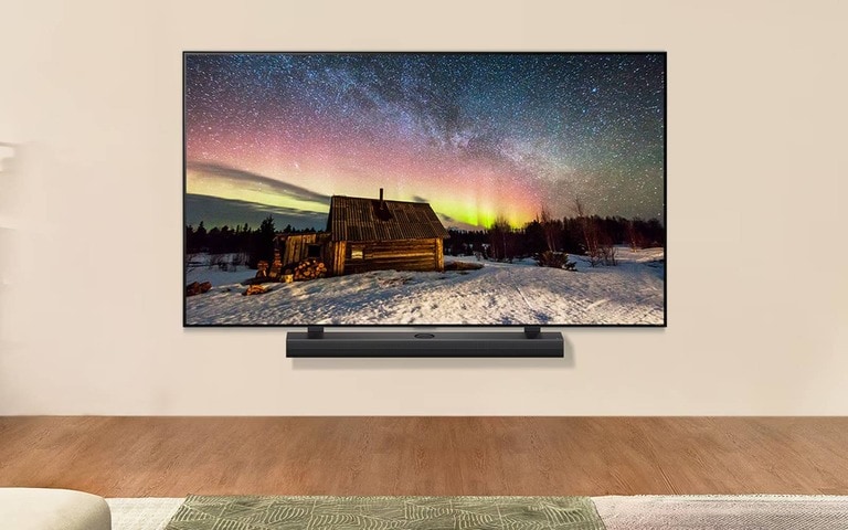 A large wall-mounted LG TV displaying a vivid night sky with stars and auroras above a rustic cabin in a snowy landscape, accompanied by a sleek soundbar below