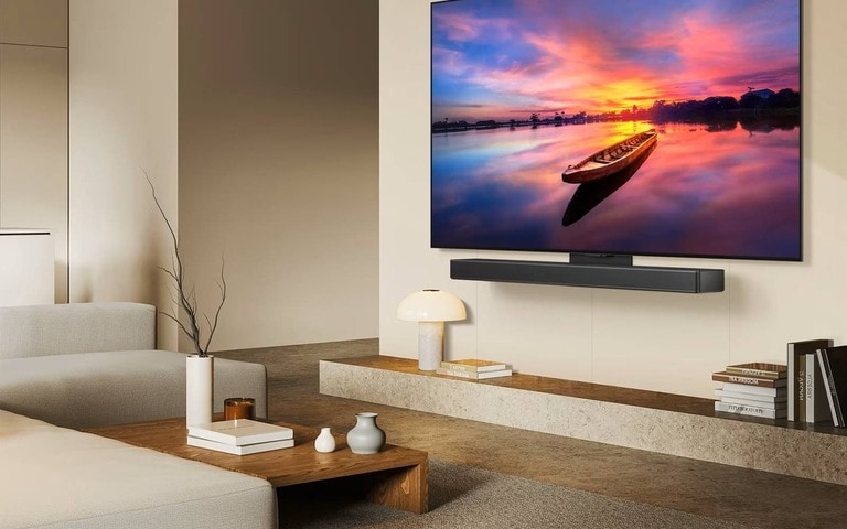 A modern living room with a large wall-mounted TV showing a vivid sunset over a calm lake, cozy beige furniture, a marble coffee table with a plant, and soft natural light filling the space.