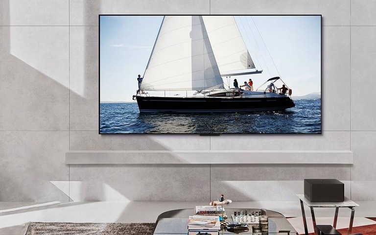 A sleek LG TV mounted on a modern living room wall, displaying a high-definition image of a sailboat gliding on the ocean, with minimalist decor and a small table featuring books and a decorative object nearby.