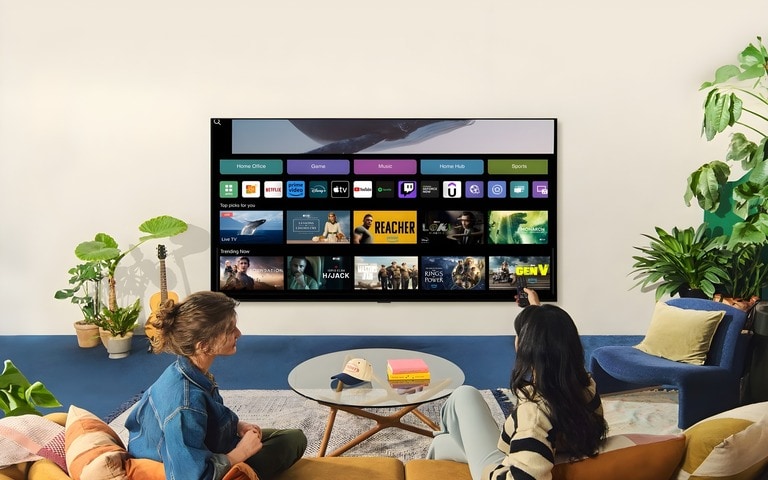 Two people sitting in a cozy living room, watching an LG smart TV displaying a user interface with streaming apps, surrounded by plants and modern decor