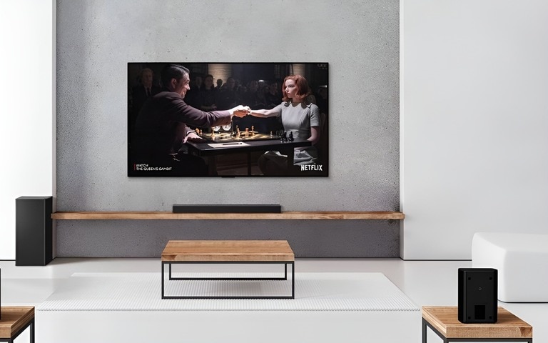 A minimalist living room with an LG soundbar and subwoofer setup under a mounted TV. The TV screen displays a Netflix show, with two rear speakers positioned for immersive sound.