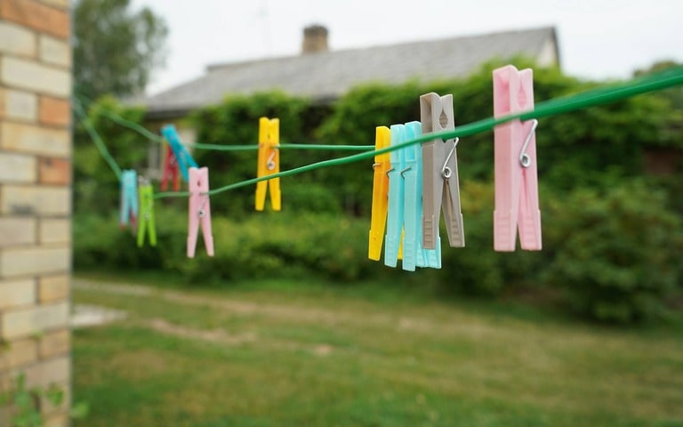 A clothesline with colorful clothespins hanging outdoors in a garden setting, with a blurred background of greenery and a house