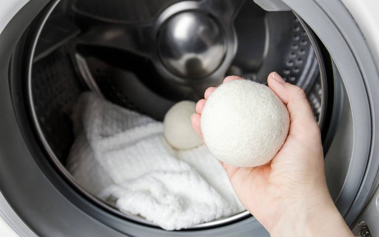 The inside of an LG washing machine or dryer with a person holding a wool dryer ball in their hand. Inside the drum, there is a white towel along with another dryer ball, emphasising the use of eco-friendly alternatives for laundry.