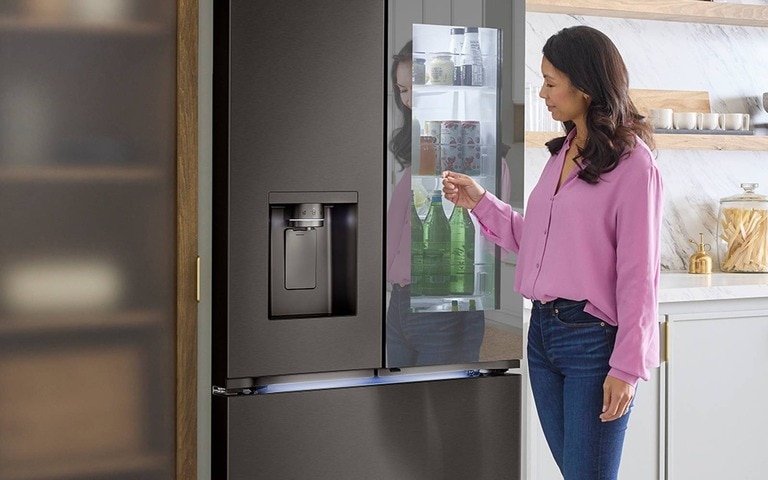 A woman interacting with a modern stainless steel LG French door refrigerator featuring a "door-in-door" design. She is accessing the easy-access compartment where drinks and frequently used items are stored, while the refrigerator also features a built-in water and ice dispenser on the left door.