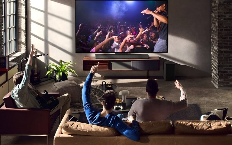 A loft-style living room with a group of friends watching a live concert on a large TV paired with an LG soundbar and subwoofer. The ambiance is cozy and vibrant, highlighting the surround sound experience.