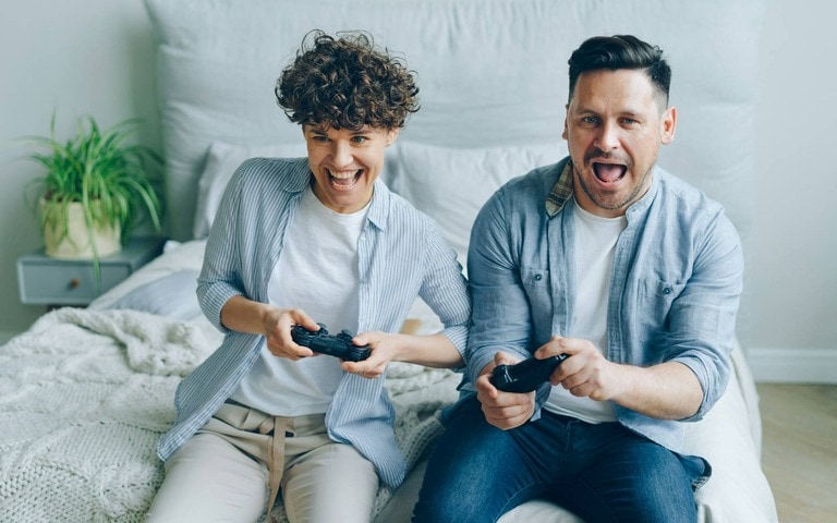 A cheerful couple sitting on a bed, enthusiastically playing video games with controllers, enjoying a relaxed and fun moment