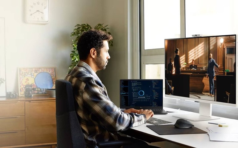 Person working at a desk with an LG monitor and a laptop. The monitor displays a scene from a video or movie, while the laptop shows coding or data. The workspace is bright and organised, with plants, artwork, and a window providing natural light.