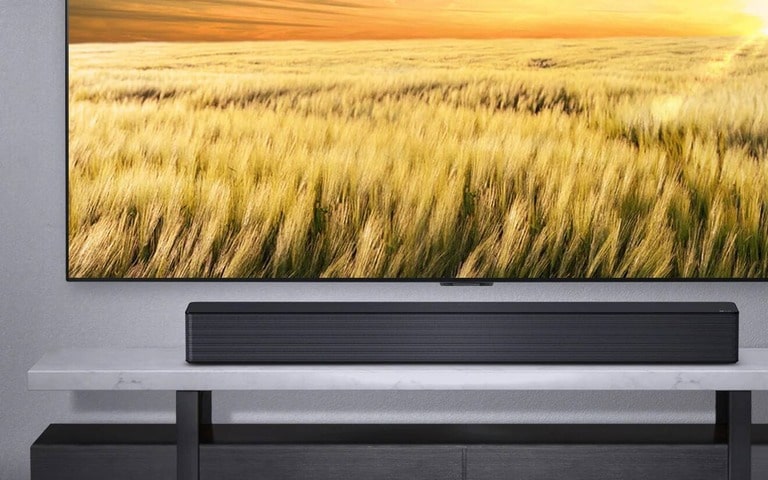 An LG soundbar placed beneath a large-screen TV displaying a serene field of golden wheat. The elegant marble-top TV console complements the sleek design of the soundbar