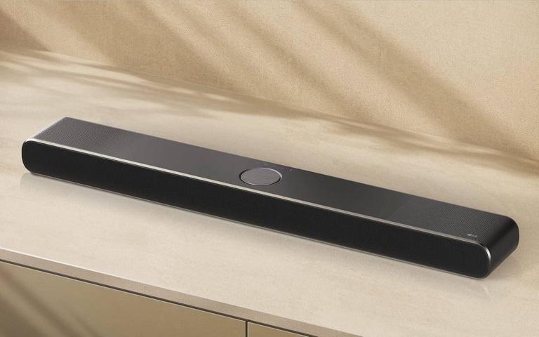 The image shows a sleek, black LG soundbar with a minimalist design, rounded edges, and perforated speaker grills on both ends, placed on a beige surface.