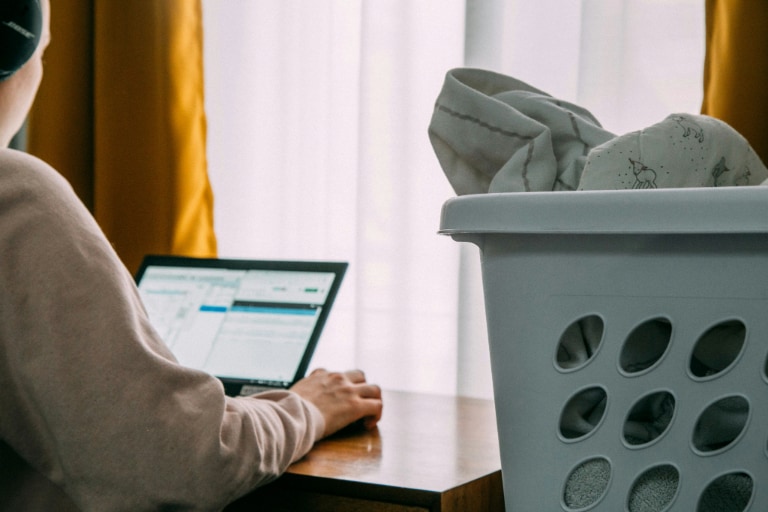 Person using a laptop next to a window with yellow curtains, while a laundry basket filled with clothes is in the foreground