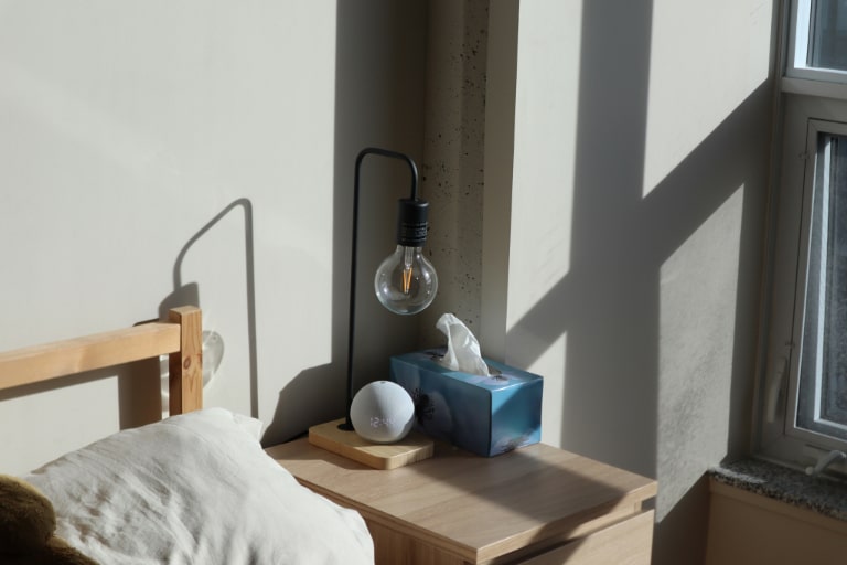A nightstand with a smart speaker in a bedroom with morning light casting shadows on a bare wall