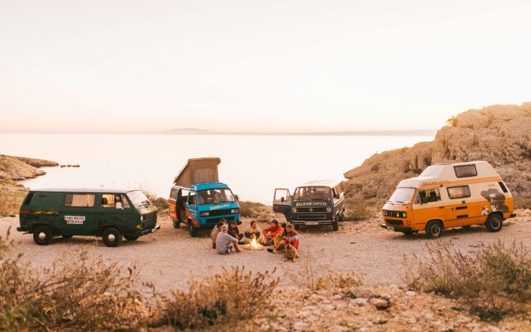 Group of people relaxing by a campfire near three colourful camper vans parked by a seaside at sunset