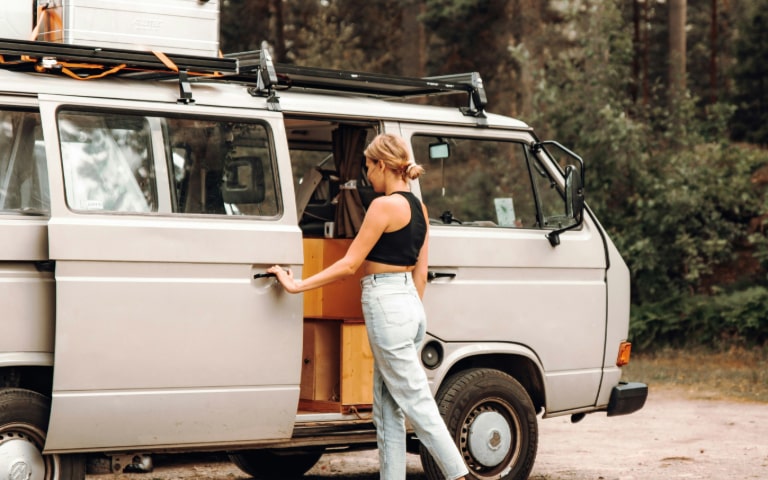 A woman opening the sliding door of a vintage camper van parked in a forest
